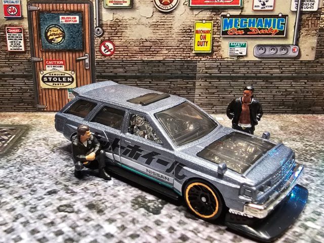 Der Nissan Maxima Drift Car aus dem 2025 Nightburnerz 5-Pack ist genau diese Art von Casting, die spaltet – und genau deshalb funktioniert’s. Metalflake Gunmetal Blue, Side-Text in Japan-Style, teal Accent wie Neon unter Streetlights… Das Ding sieht aus wie ein “Daily”, der nachts heimlich Angle-Training macht. Und die Crew? Steht schon bereit: einer hockt am Schweller, einer checkt den Look – alles wie immer vor dem ersten Slide.
Specs & Vibes:
Brand: Hot Wheels
Modell: Nissan Maxima Drift Car
Serie: 2025 Nightburnerz 5-Pack
Farbe: Metalflake Gunmetal Blue
Side-Deco: White Stripe mit “Nissan”, teal Hot Wheels Logo & Stripe, schwarzer japanischer Schriftzug
Vibe: Sleeper-Body, Drift-Soul – 1:64 Street Spec
Wenn der Winkel stimmt, ist alles andere egal.