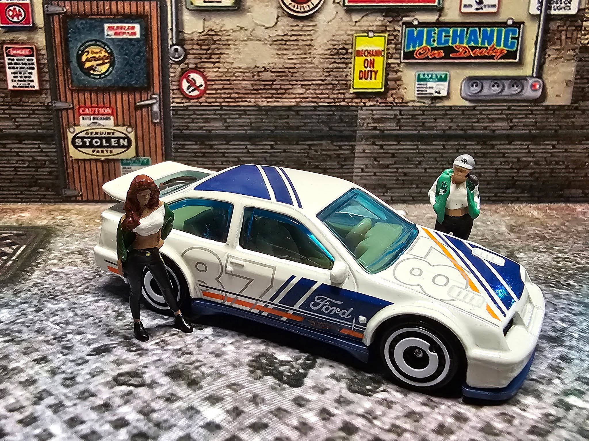 🏁 Box-Flares, Boost & 80s Attitude.
Der ’87 Ford Sierra Cosworth ist nicht einfach “Retro” – er ist die Ära, in der Turbos noch respektlos waren. In Weiß mit Dark-Blue Stripes und dem fetten „87“ wirkt er wie ein Touring-Car, das sich in die Stadt verirrt hat… und dort sofort die Regeln übernimmt. Die Crew steht rum wie bei ’nem illegalen Setup-Check: Wer dreht als Erstes den Ladedruck hoch?
Specs & Vibes:
Brand: Hot Wheels
Modell: ’87 Ford Sierra Cosworth
Serie: 2025 Nightburnerz 5-Pack
Farbe: White
Deco: Dark Blue Stripes (Haube/Dach/Seiten), Orange Stripe, graues „87“ (Haube & Seiten)
Logos: White Ford, orange Hot Wheels, orange „LWC Racing“
Vibe: Street-legal? Vielleicht. Street-royalty? Safe.
Wenn der Boost kommt, wird die Nostalgie zur Waffe.